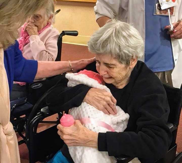 woman places hand on shoulder of woman sitting down holding swaddled baby doll and baby bottle in hand