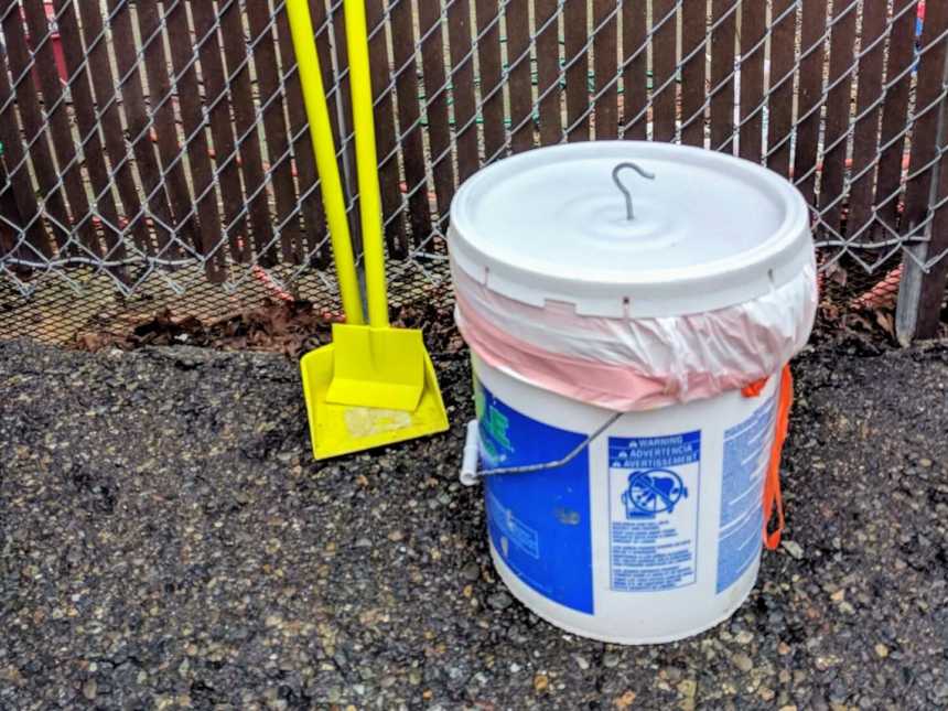 a pooper scooper leaned against a wire fence with a white paint bucket next to it