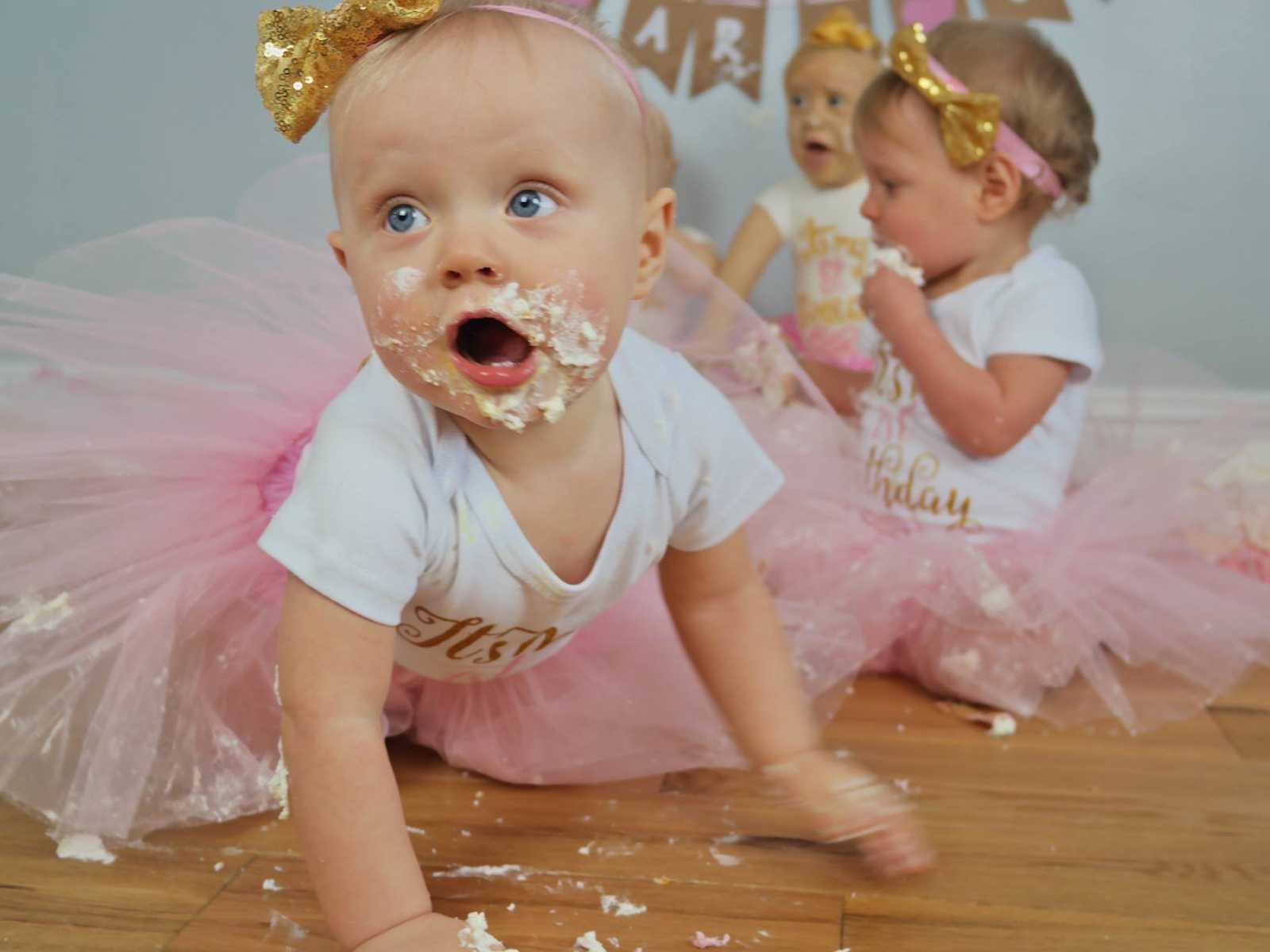 infant girl crawling on ground with cake all over her face with another infant eating cake in background