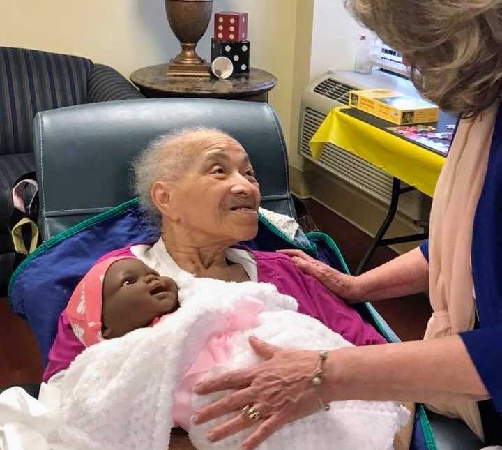 elderly woman smiling up at woman standing over her while holding baby doll in arms that is swaddled in pink blanket
