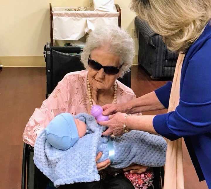 alzheimer patient with sunglasses on holds swaddled blanket in blue blanket while woman offers purple baby bottle