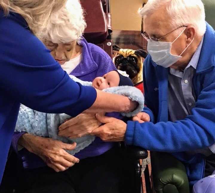 woman hands alzheimer patient a baby doll who is sitting next to a an elderly man with mask on and stuffed dog