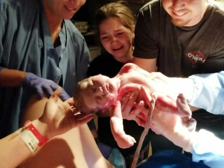 newborn baby with umbilical chord still attached is held in air by nurses as father looks down smiling and older sister cries