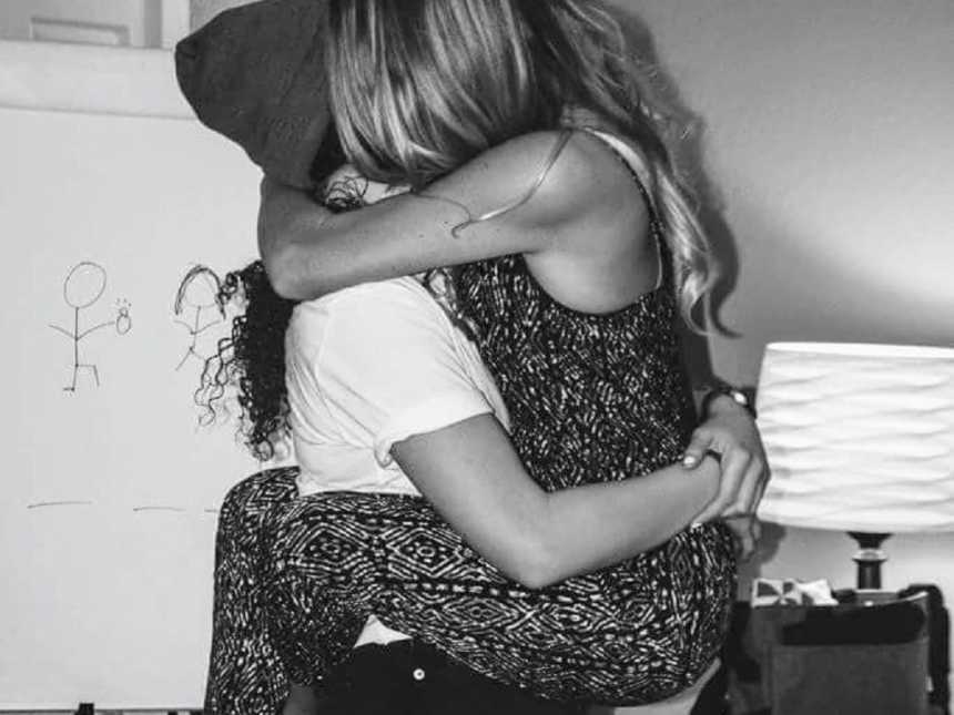 women holding up another woman in her arms with whiteboard in background with doodle of person proposing 