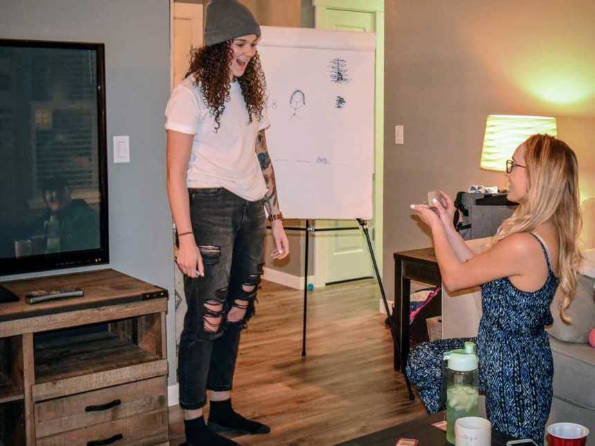 blonde-haired woman proposing in living room to brown curly-haired girlfriend standing next to a whiteboard with doodles