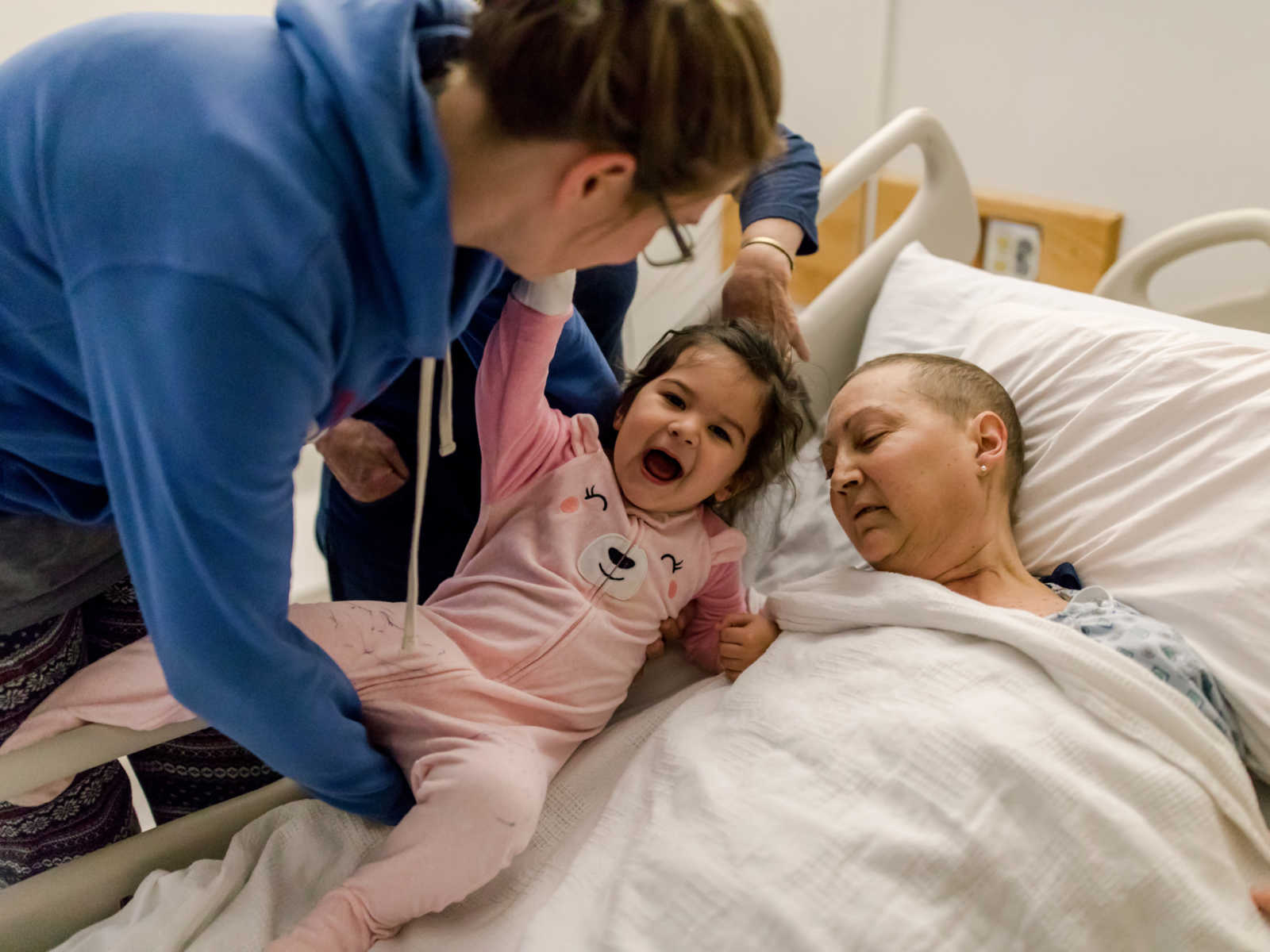 Toddle in pink onesie is smiling while being placed in hospital bed next to woman with non-hodgkin's lymphoma