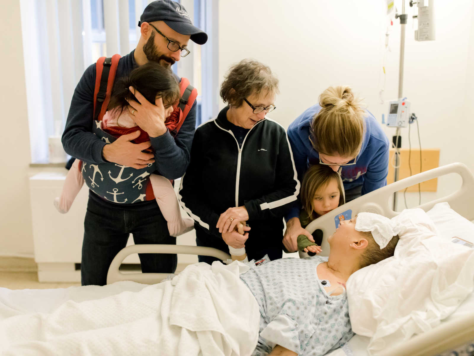 Sufferer of non-hodgkin's lymphoma lies in bed holding mothers hand and looks up at sister, niece, nephew, and brother in law