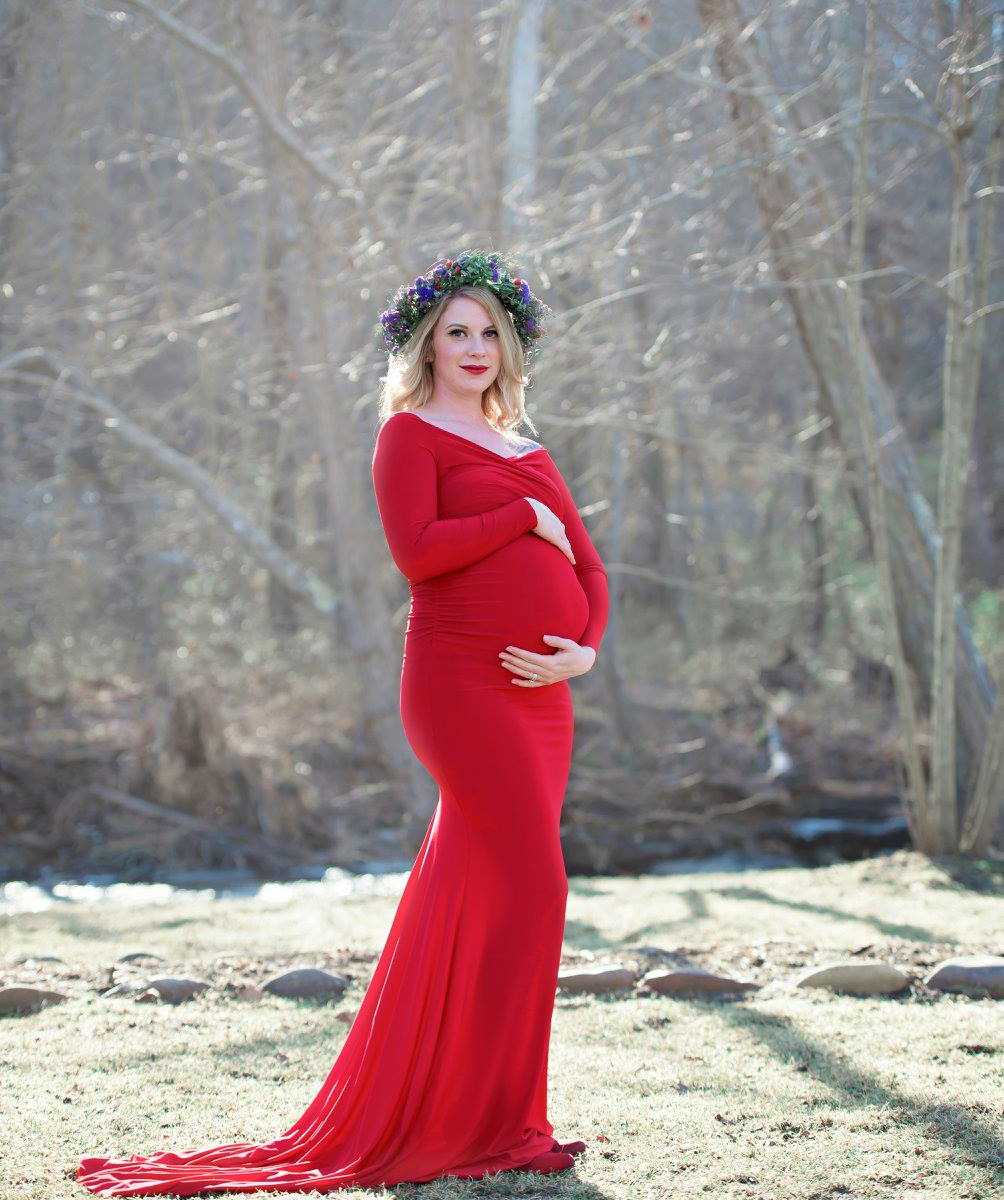 pregnant widower in red dress and flower crown holds baby with trees in the background