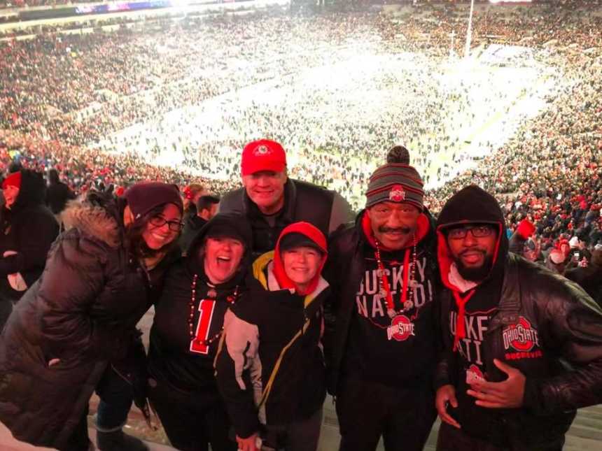 six people smiling in ohio state gear with a packed stadium and people rushing the field in the background