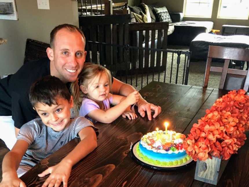 Father who lost preemie daughter crouches down next to son and daughter who sit at table with birthday cake