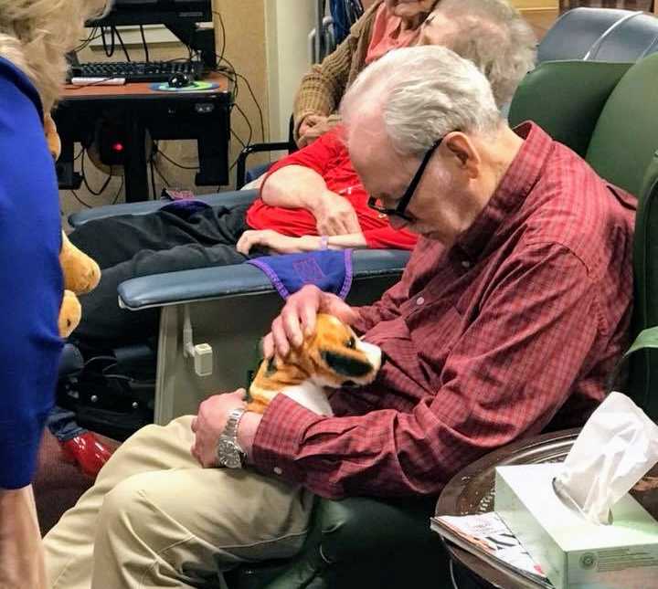 elderly man with black glasses and red shirt looks down at stuffed animal dog in his lap
