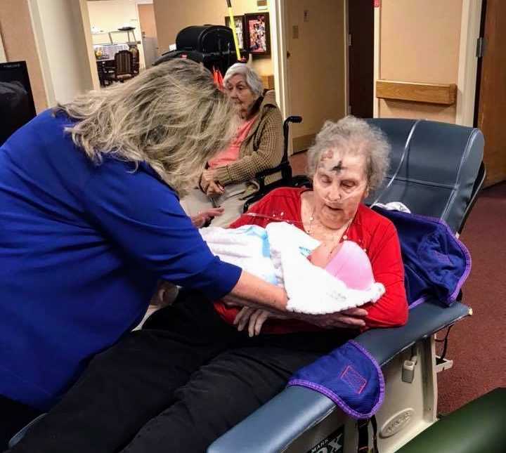 woman hands baby doll to elderly woman in reclined chair who has an as cross on forehead and oxygen up her nose