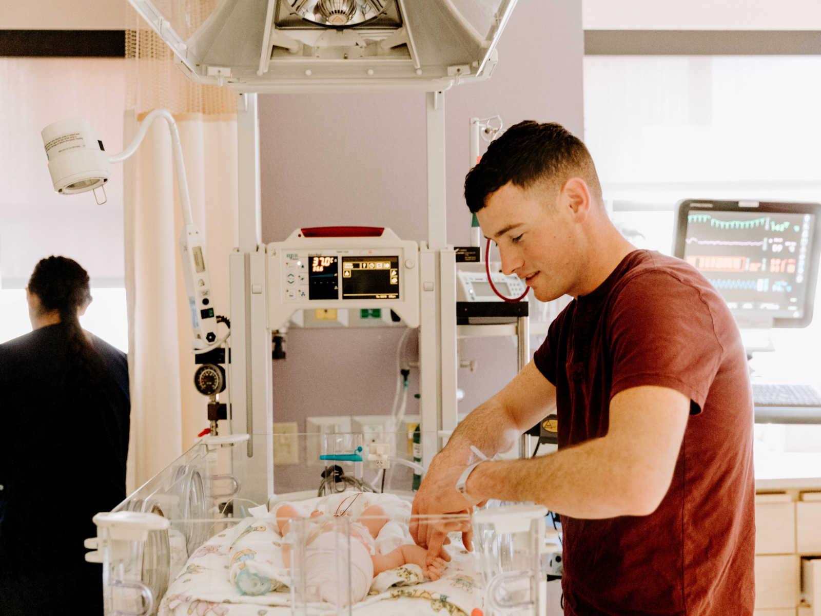 Father stands over newborn baby and holds it's hand in NICU