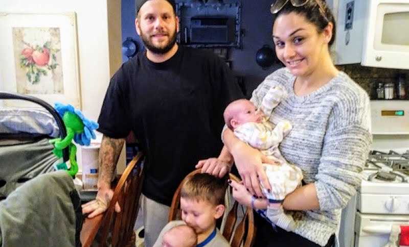 Brother who donated bone marrow to twin siblings holds baby in lap while mom holds the other next to husband