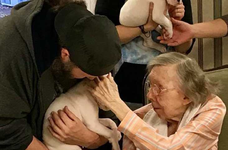 man leans over with white puppy in his arms to let elderly woman pet it