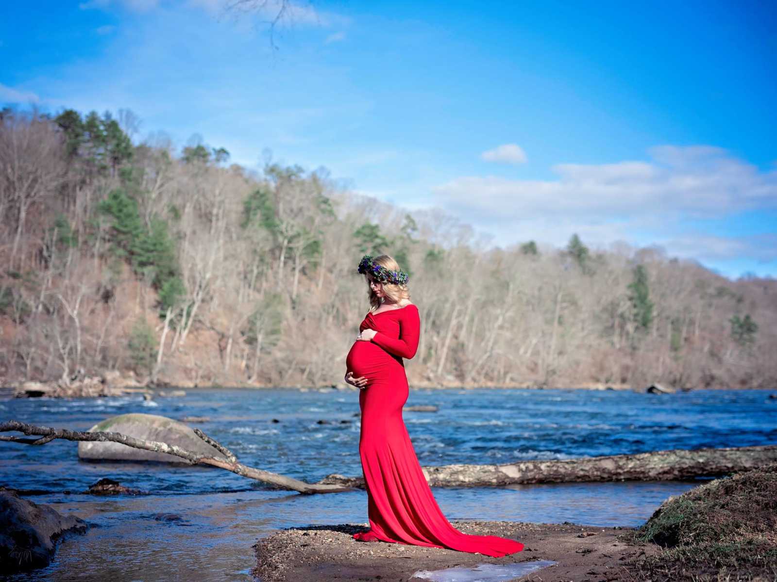pregnant widower in red dress and flower crown holds stomach on shore of river