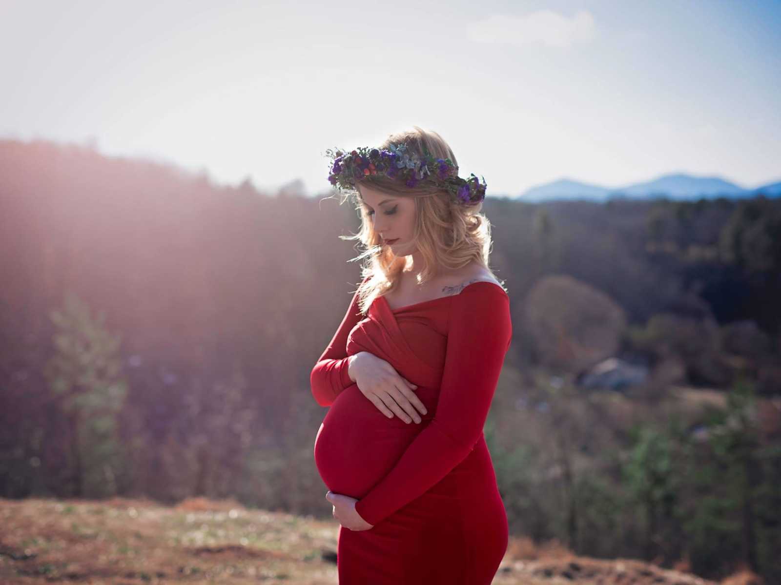 pregnant widower in red dress and flower crown holds stomach on hill with forest in background