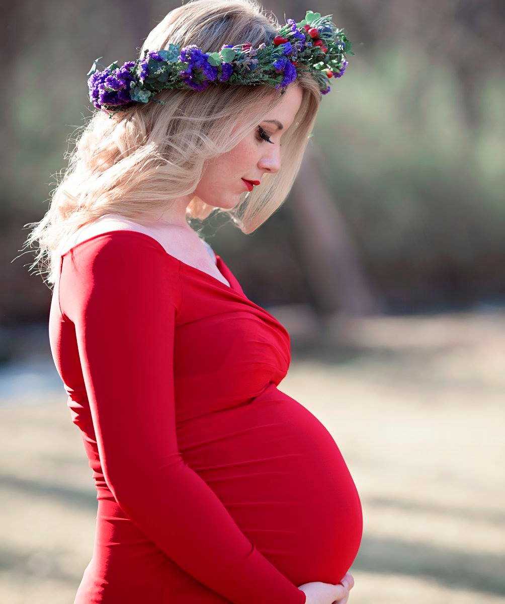 pregnant widower in red dress and flower crown looks down at her stomach while holding it