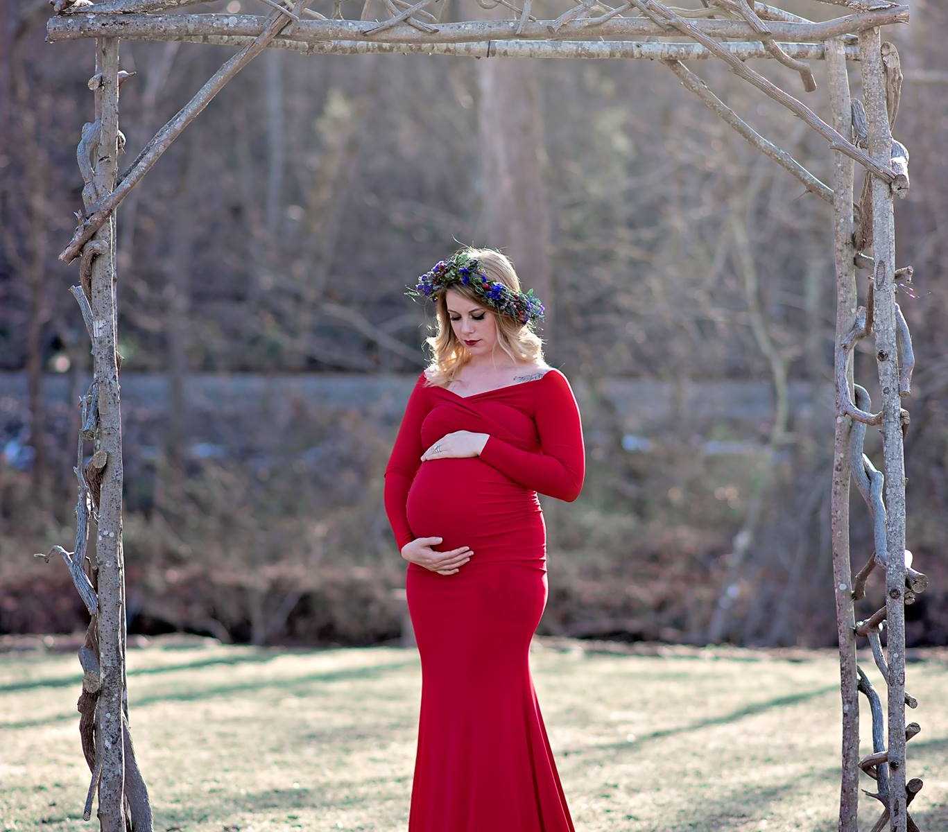 pregnant widower in red dress wearing a flower crown holds her stomach under wooden arch