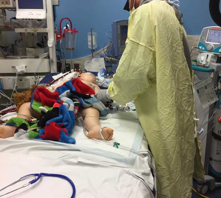 person leaning over side of hospital bed in yellow hospital gown looking over toddler boy lying in hospital bed