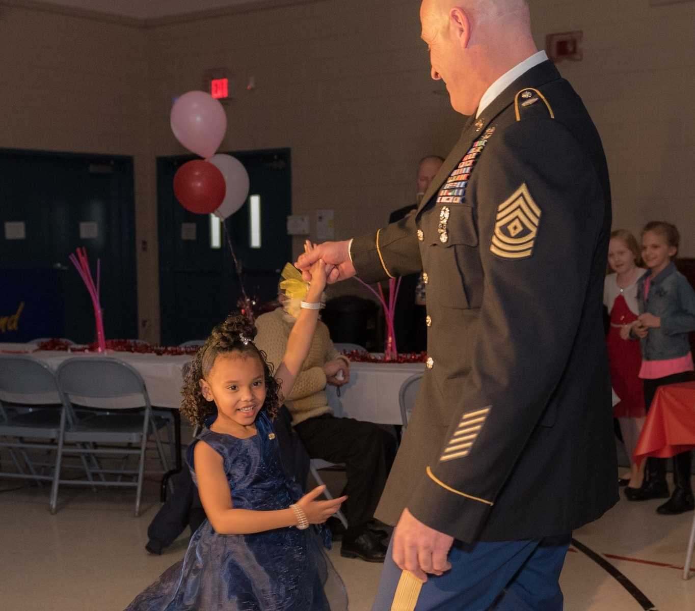 national guard soldier twirling little girl in gymnasium with table and pink balloons in background