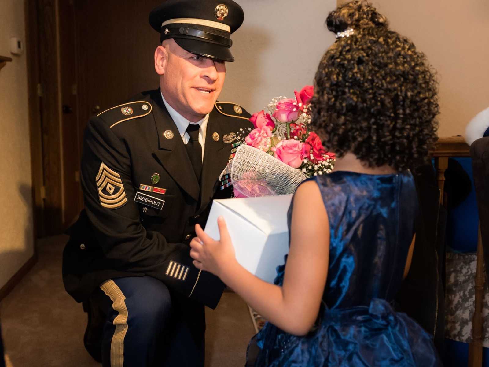national guard soldier kneeling with bouquet of pink roses in front of little girl in fancy blue dress with curly brown hair
