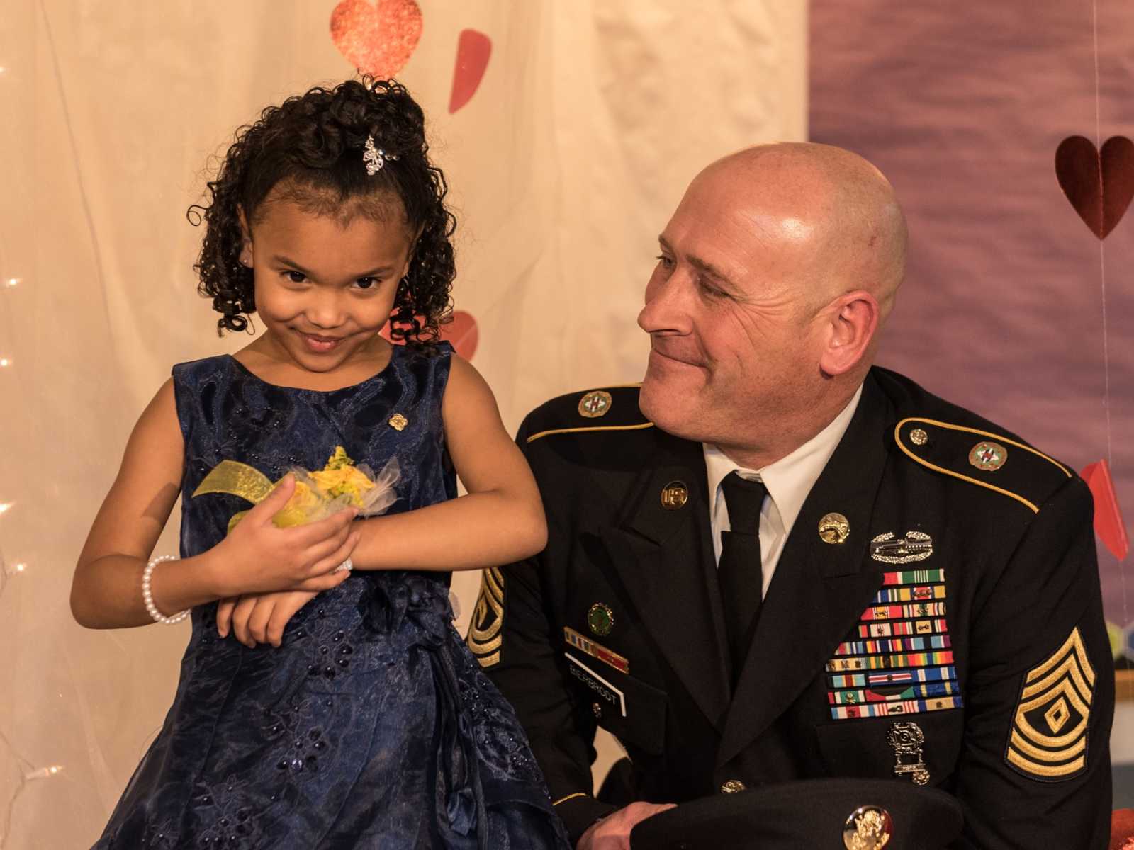 little girl in fancy blue dress holding yellow flower wristlet next to soldier smiling at her in front of heart backdrop