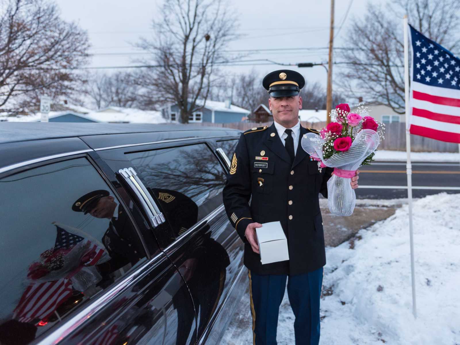 national guard soldier standing in snowy driveway next to limo and american flag holding bouquet of pink roses and white box