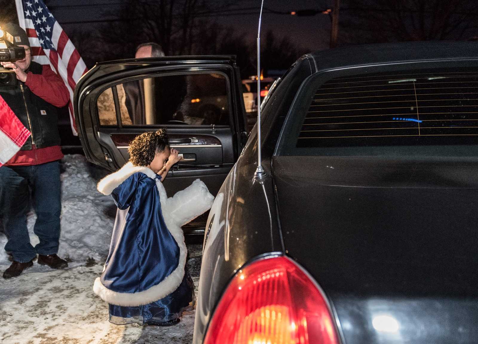 Little girl with curly brown hair in blue velvet cape getting into limo with cameraman filming with american flag in hand