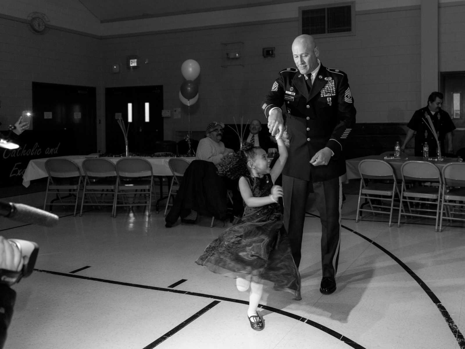 soldier twirling daughter in gymnasium with people in background watching in black and white