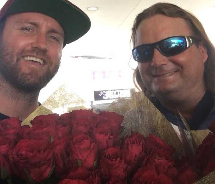 homeless man and bearded man with baseball hat on smile in selfie with bouquet of red roses