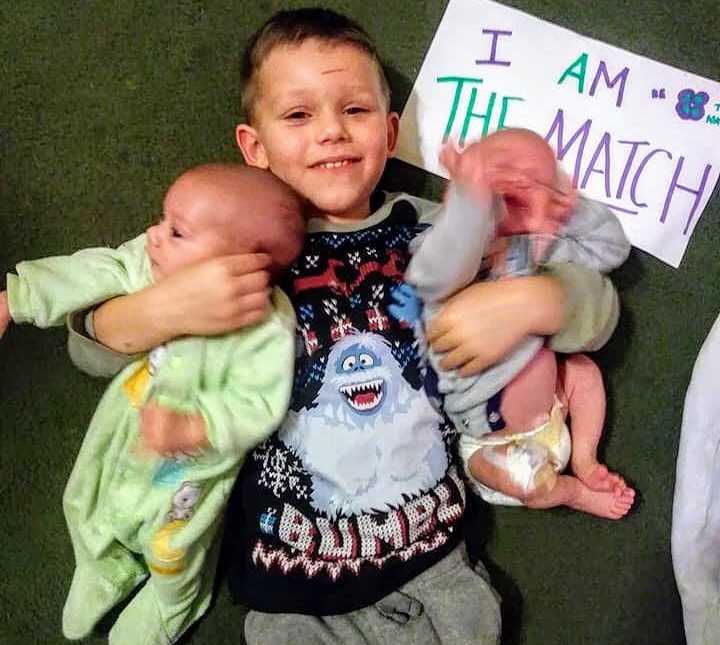 Brother who donates bone marrow to baby siblings lies on floor with them in his arm next to sign saying, "I am the match"
