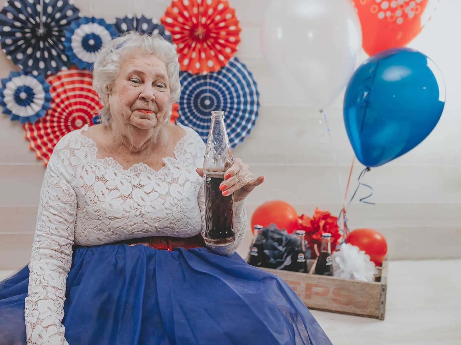 elderly woman in white lace top red belt and blue skirt smiles holding a glass bottle of soda next to wooden crate of pepsi