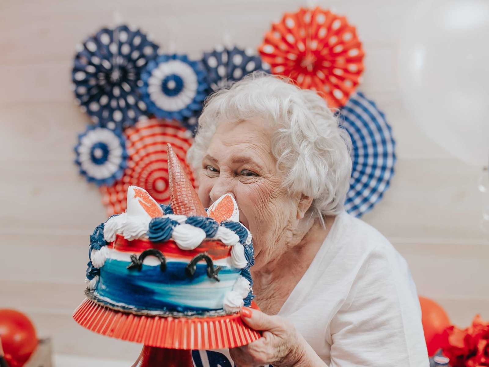 elderly woman taking bite out of red white and blue cake that resembles a unicorn