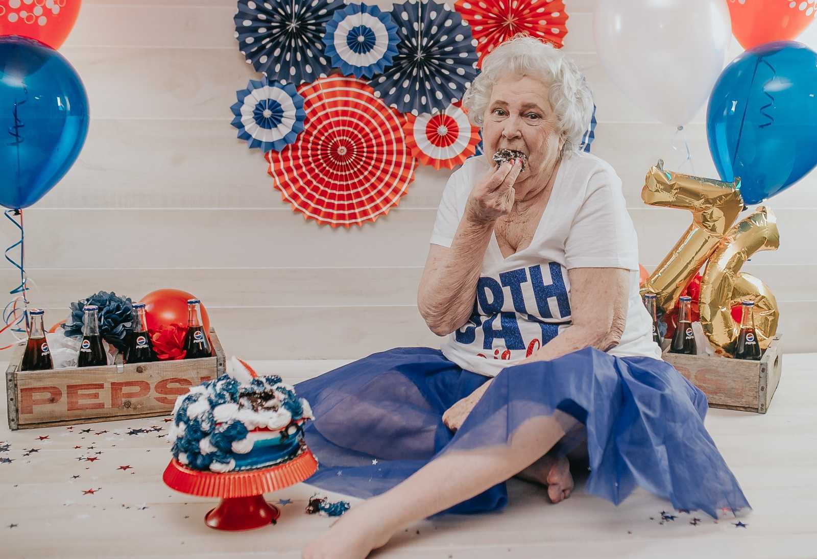elderly woman sits on floor next to red white and blue cake with handful of cake close to her mouth