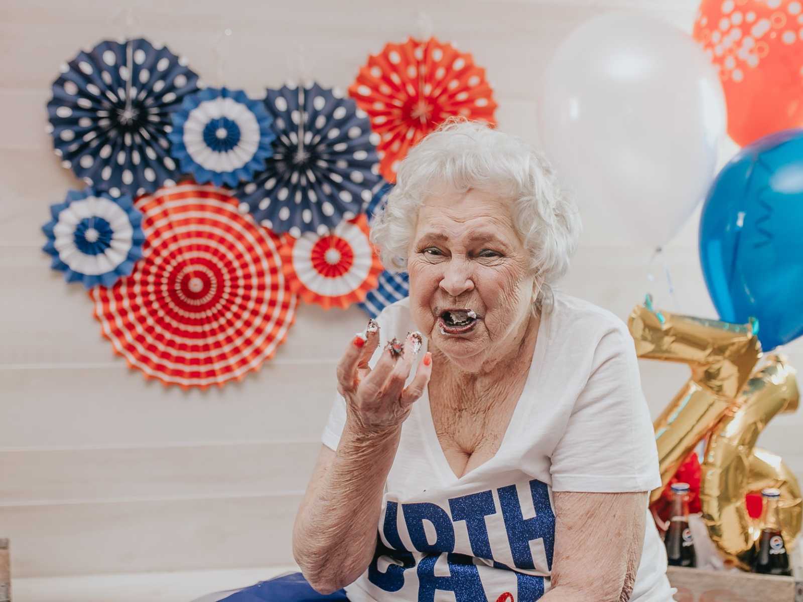 elderly woman eating cake with frosting all over her hands and red white and blue decorations in the background