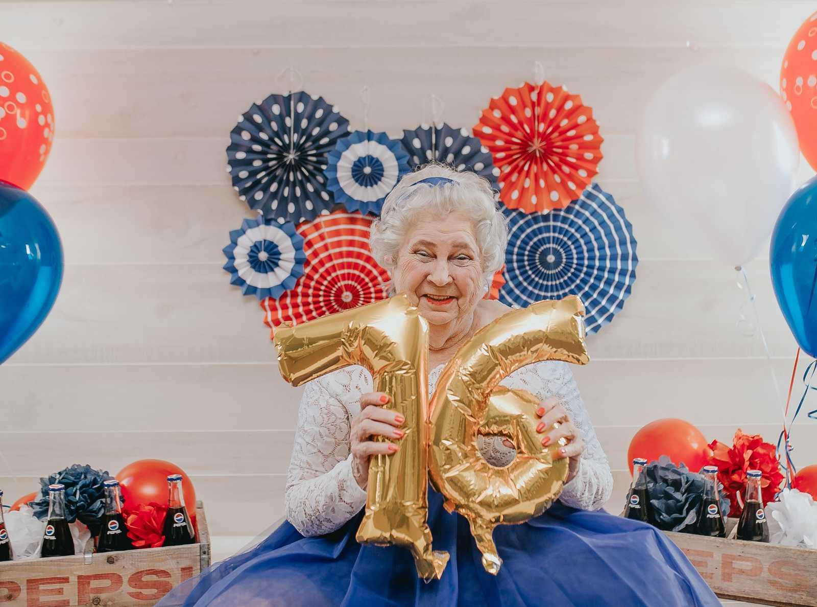 elderly woman holding up gold balloons of number 76 next to crates of pepsi and red white and blue decorations