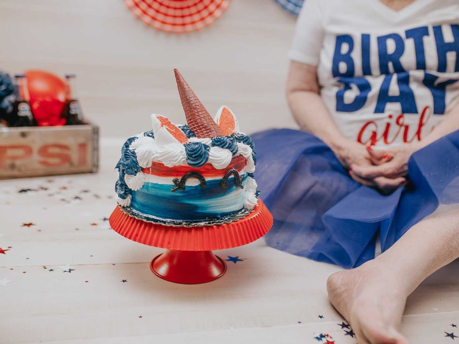 close up of red white and blue unicorn cake with elderly woman sitting on ground next to it