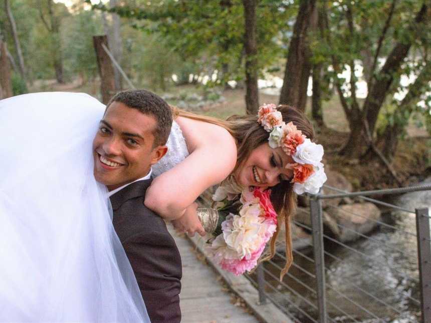 groom smiles while holding bride over shoulder on bridge over body of water