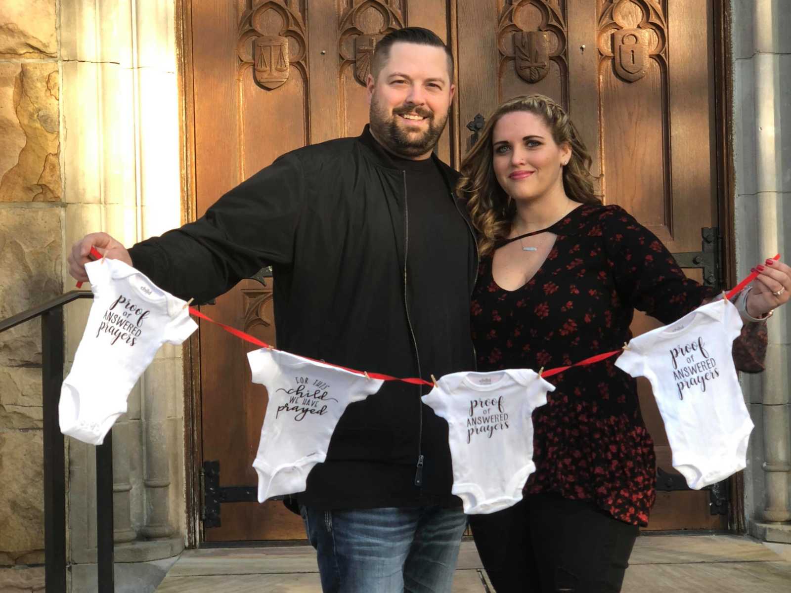 husband and wife smile while holding up a string that has four baby onesies attached
