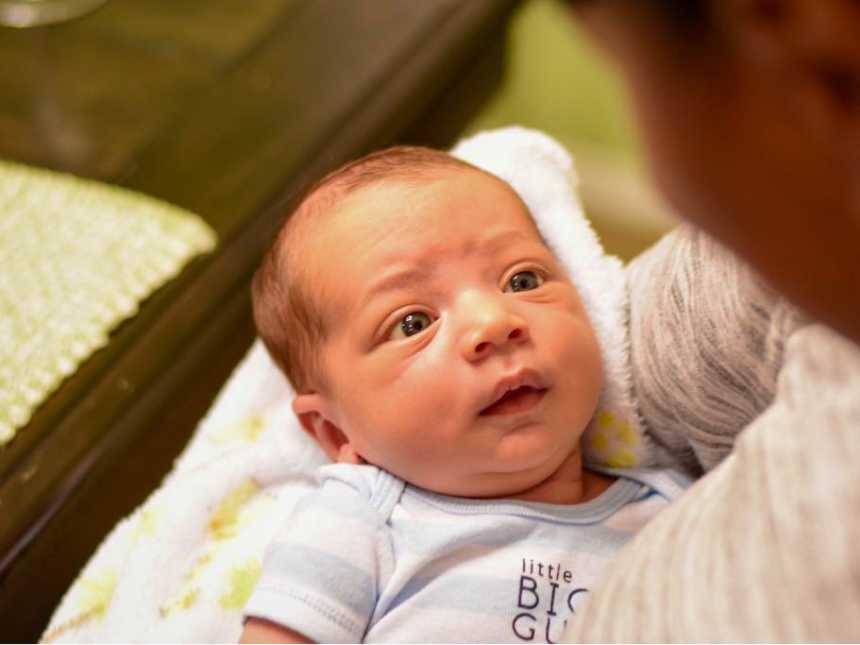 newborn baby in blue and white striped onesie that says, "little big guy" staring up at a woman