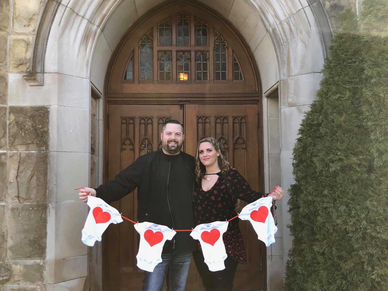 husband and wife stand in front of church holding up string of four baby onesies with red hearts on them