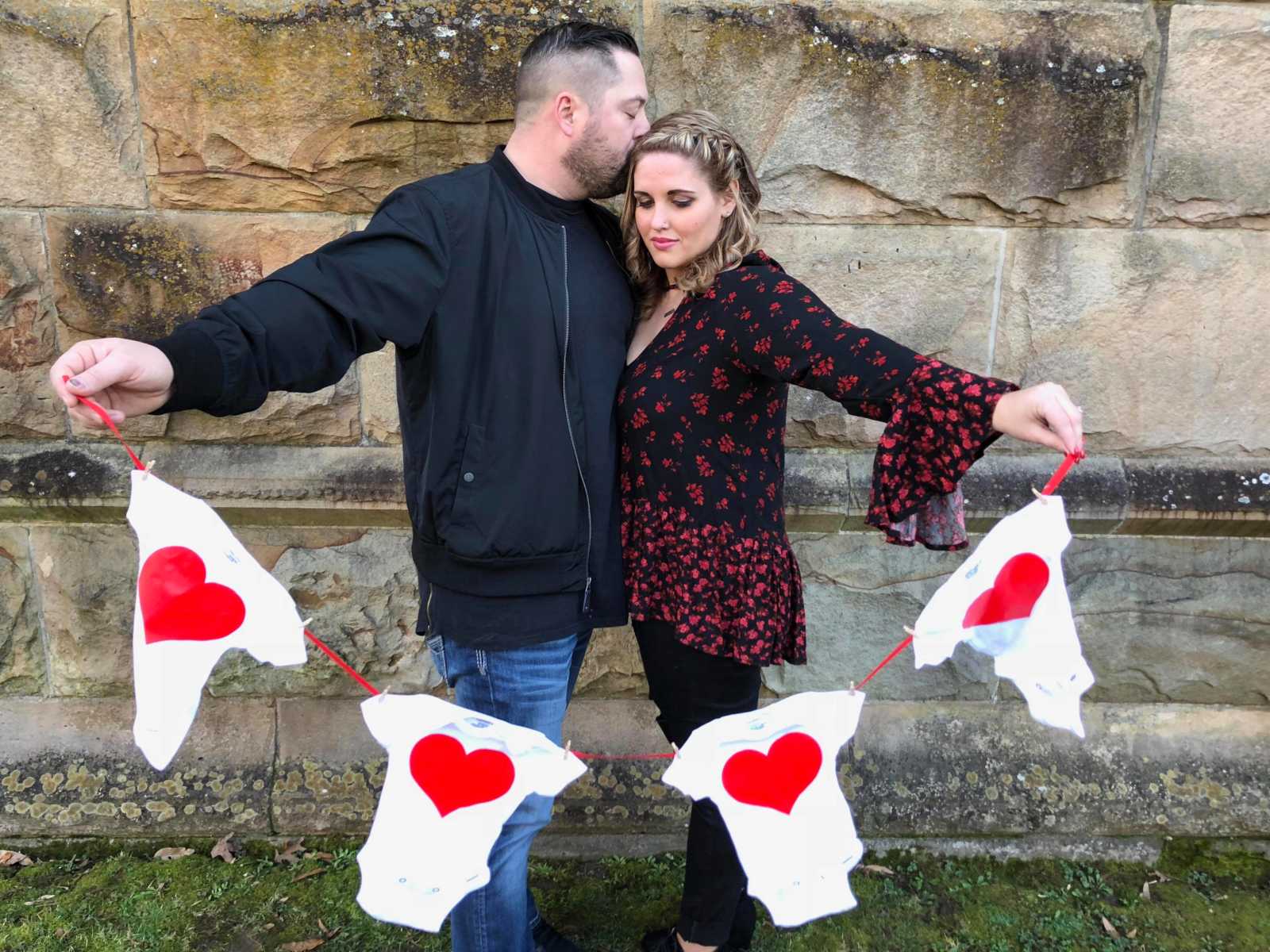 husband kisses wife on forehead as they hold up string of four baby onesies with red hearts on them