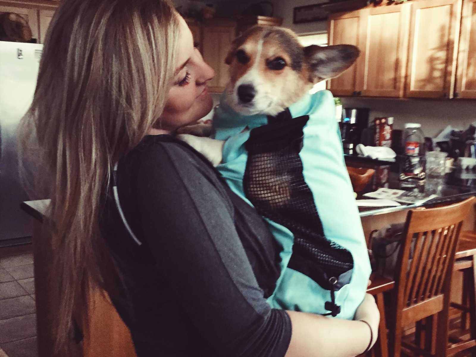 blonde woman holds dog in blue drawstring bag to her chest in kitchen