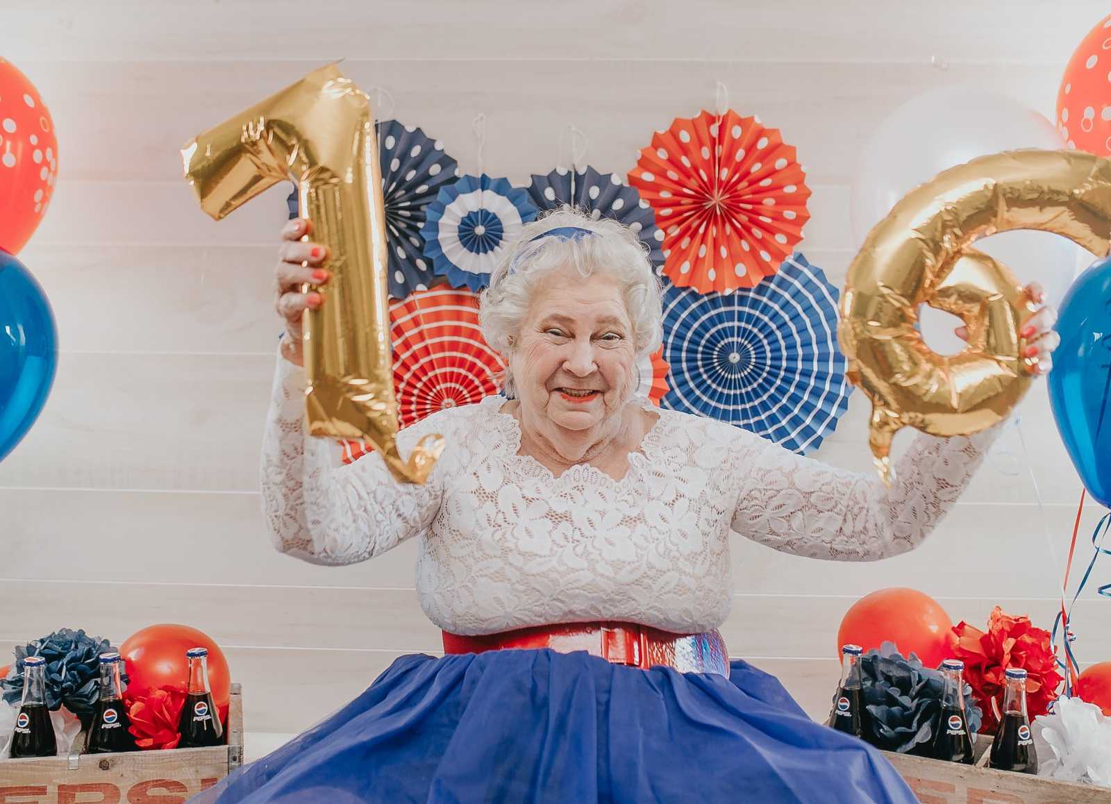 elderly woman smiling while holding up a gold 7 and 6 balloon on either side of her