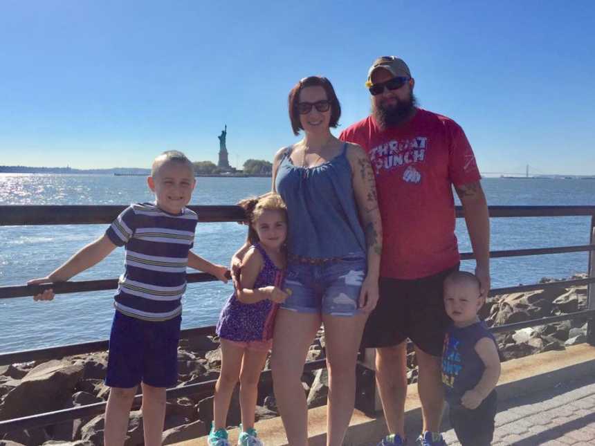 husband and wife stand next to their two sons and daughter smiling with statue of liberty in background