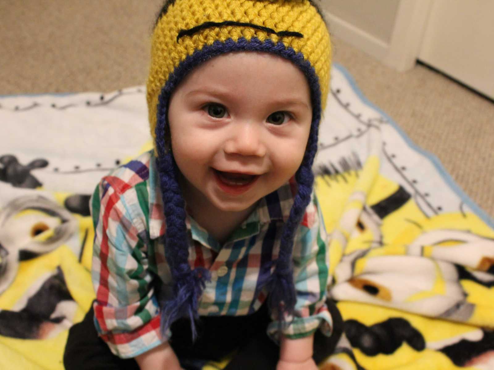 infant boy sits on minion blanket while looking up smiling with yellow knit hat on and plaid button down shirt