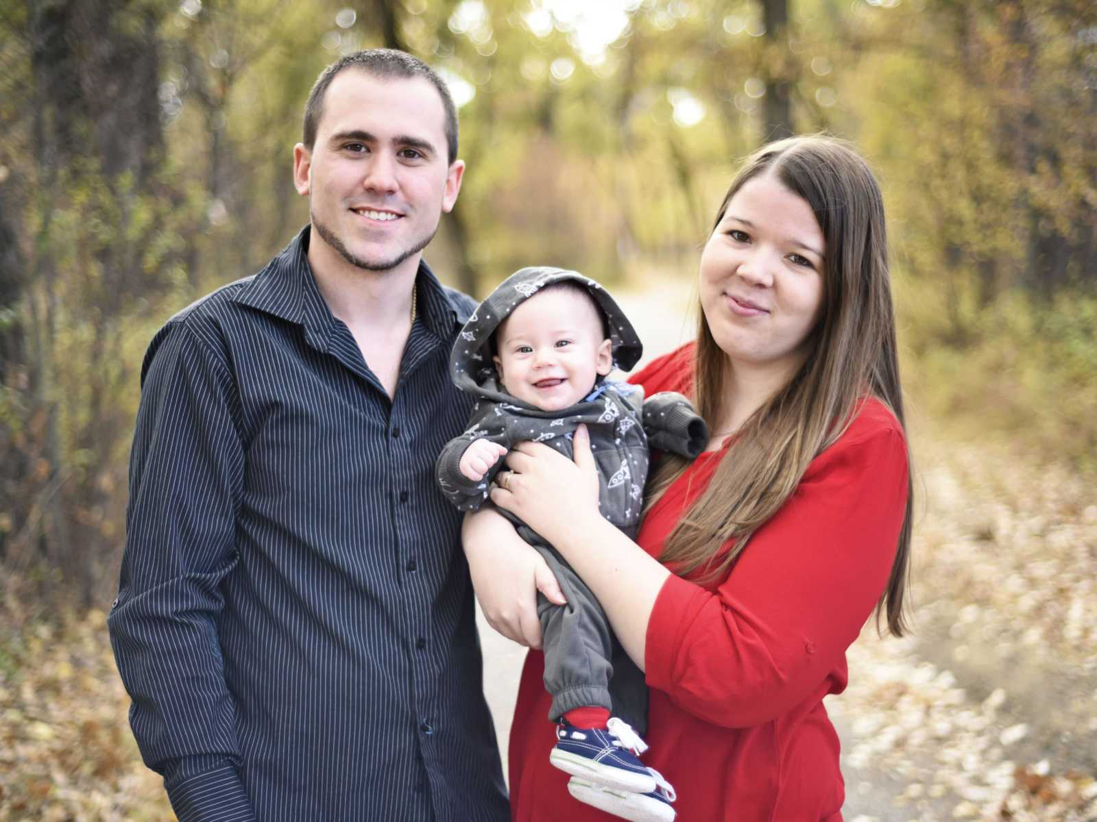 mother smiles standing with baby in her arms next to husband on trail in woods