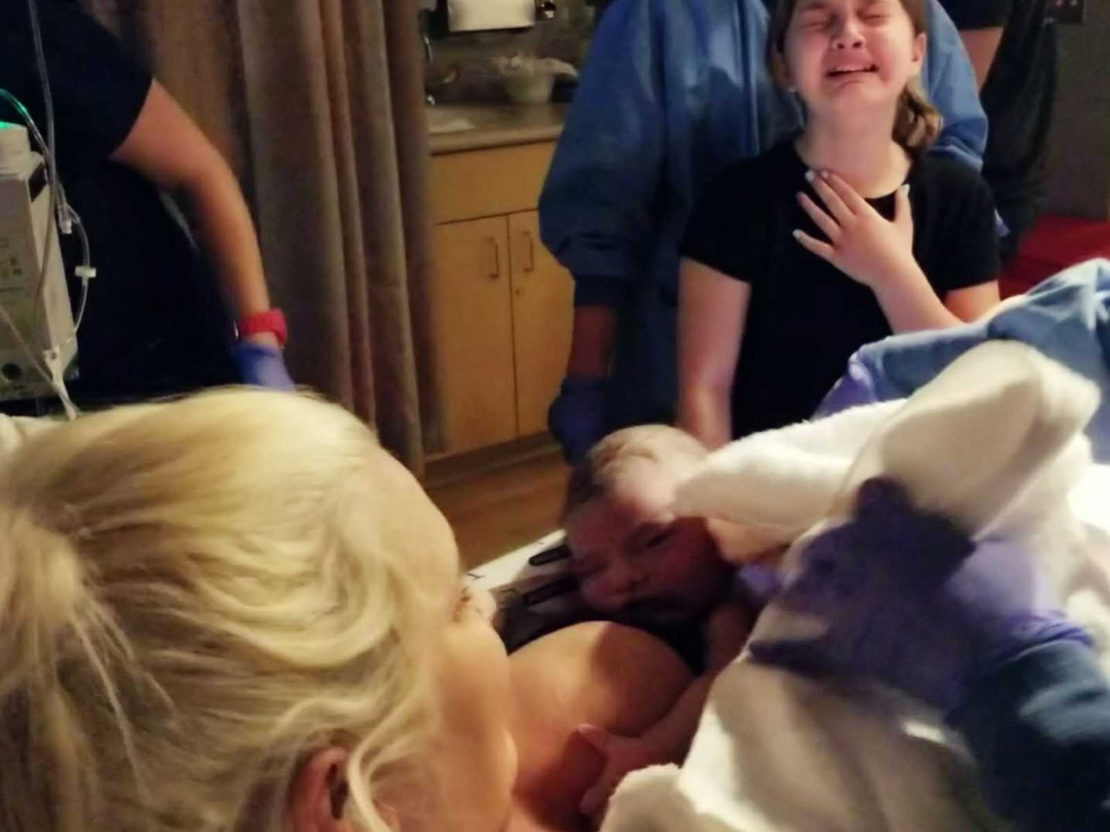 older sister holds hand to chest as she cries at the birth of her newborn sibling held in moms arm in hospital bed
