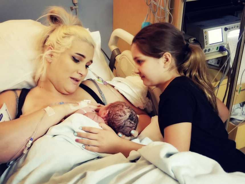 mother breastfeeds newborn in hospital bed while older daughter has hand on baby's back